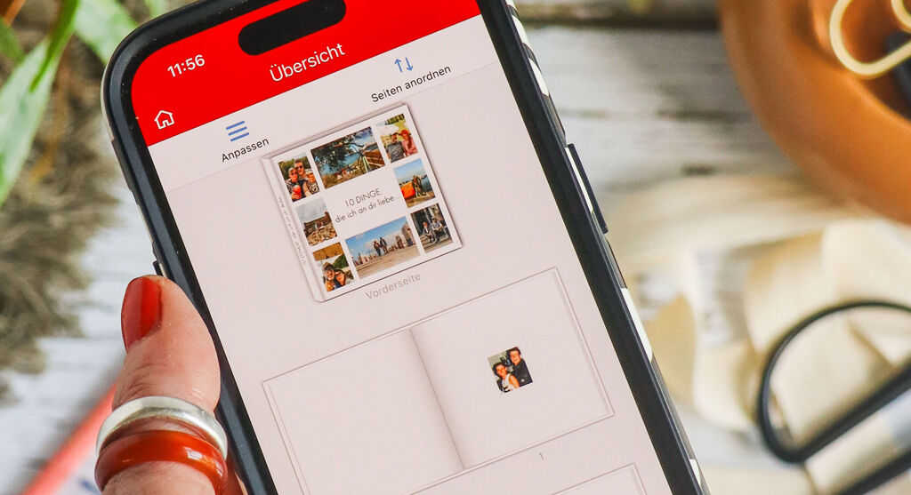 A kép előterében egy pirosra lakkozott körmű női kéz tart egy okostelefont. A kijelzőn a mobilos CEWE FOTÓ app szerkesztőfelülete van megnyitva. A korábban bemutatott fotókönyvről van szó, amely a kép elmosódott hátterében, az asztal felületén látható. Szintén elmosódva kivehető egy zöld növény, valamint kreatív kellékek, például színes dekortapasztekercsek, tollak, olló és ajándékszalag.